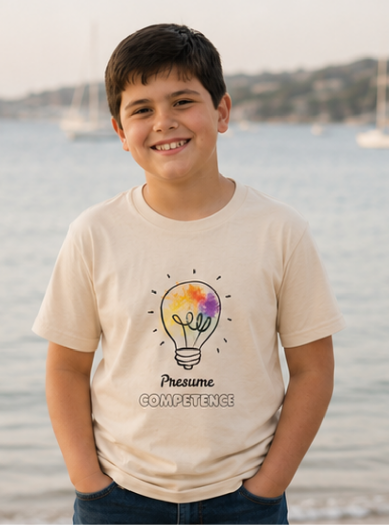 A smiling child wears Presume Competence T-shirt in a soft natural cream color, featuring a colorful light bulb illustration and the empowering message. Presume Competence. Set against a calm outdoor background, the image reflects confidence, kindness, and inclusion while celebrating the unique strengths of every child.
This meaningful graphic tee is designed for children of all abilities.